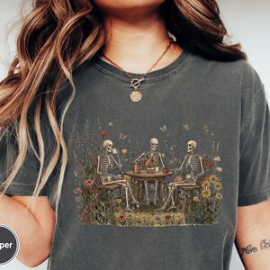 Może przedstawiać: Ciemnoszary t-shirt z kr&oacute;tkim rękawem z nadrukiem graficznym przedstawiającym trzy szkielety siedzące przy okrągłym stole na polu pełnym kolorowych polnych kwiat&oacute;w. Szkielety piją z kubk&oacute;w i nalewają płyn z dzbanka do filiżanki.