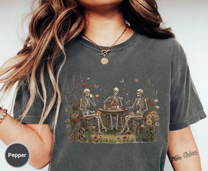 Może przedstawiać: Ciemnoszary t-shirt z kr&oacute;tkim rękawem z nadrukiem graficznym przedstawiającym trzy szkielety siedzące przy okrągłym stole na polu pełnym kolorowych polnych kwiat&oacute;w. Szkielety piją z kubk&oacute;w i nalewają płyn z dzbanka do filiżanki.