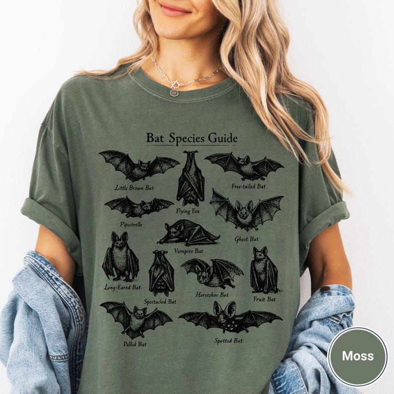 May include: Moss green t-shirt featuring a black graphic of a bat species guide. The guide includes illustrations and names of various bat species such as the Little Brown Bat, Flying Fox, and Vampire Bat. The text "Bat Species Guide" is at the top.