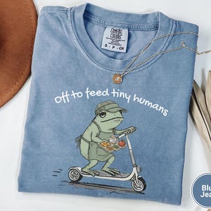 May include: Blue jean Comfort Colors t-shirt with the phrase "off to feed tiny humans." The graphic shows a green frog on a scooter carrying a tray of food. A gold necklace is draped on the shirt.