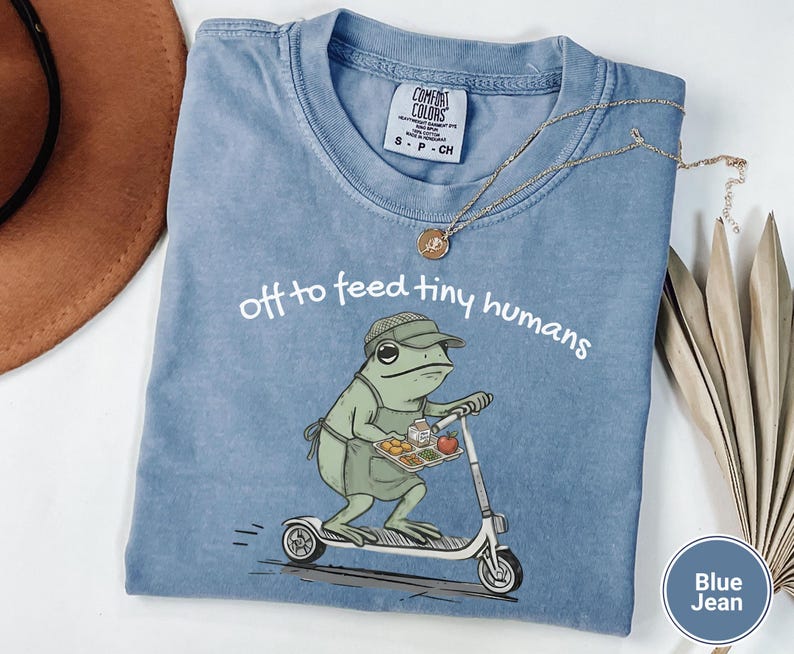 May include: Blue jean Comfort Colors t-shirt with the phrase "off to feed tiny humans." The graphic shows a green frog on a scooter carrying a tray of food. A gold necklace is draped on the shirt.
