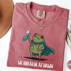 Pode incluir: Uma t-shirt de manga curta cor carmesim com o gr&aacute;fico de um sapo verde usando uma capa azul-petr&oacute;leo e um estetosc&oacute;pio, segurando um ressuscitador manual. Abaixo do sapo, o texto diz WE BREATHE AT DAWN.