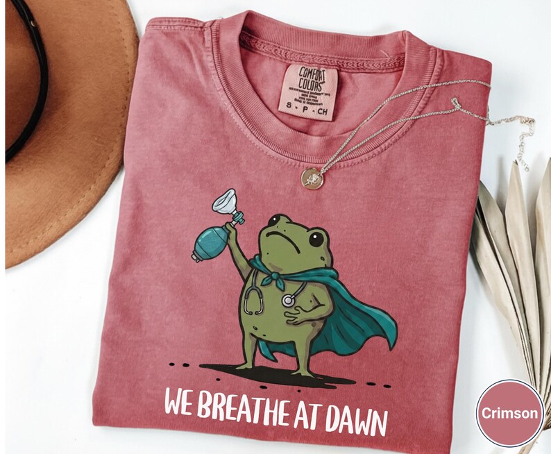 Pode incluir: Uma t-shirt de manga curta cor carmesim com o gr&aacute;fico de um sapo verde usando uma capa azul-petr&oacute;leo e um estetosc&oacute;pio, segurando um ressuscitador manual. Abaixo do sapo, o texto diz WE BREATHE AT DAWN.