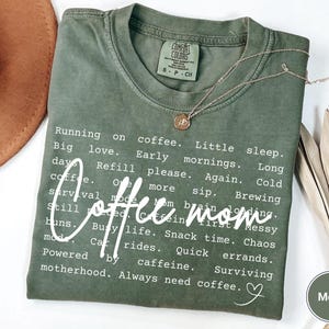 May include: A moss green Comfort Colors t-shirt with the words "Coffee Mom" in a white script font. The shirt has a list of coffee-related phrases. A gold necklace with a pendant is on the shirt.