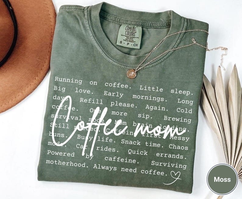 May include: A moss green Comfort Colors t-shirt with the words "Coffee Mom" in a white script font. The shirt has a list of coffee-related phrases. A gold necklace with a pendant is on the shirt.