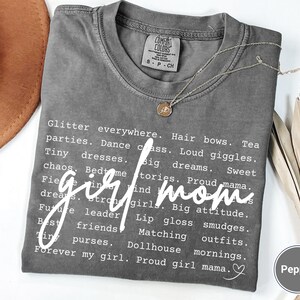 May include: A grey t-shirt with the words "girl mom" in a cursive font. The shirt features a list of related words in a white font. A gold necklace with a pendant is on the shirt.