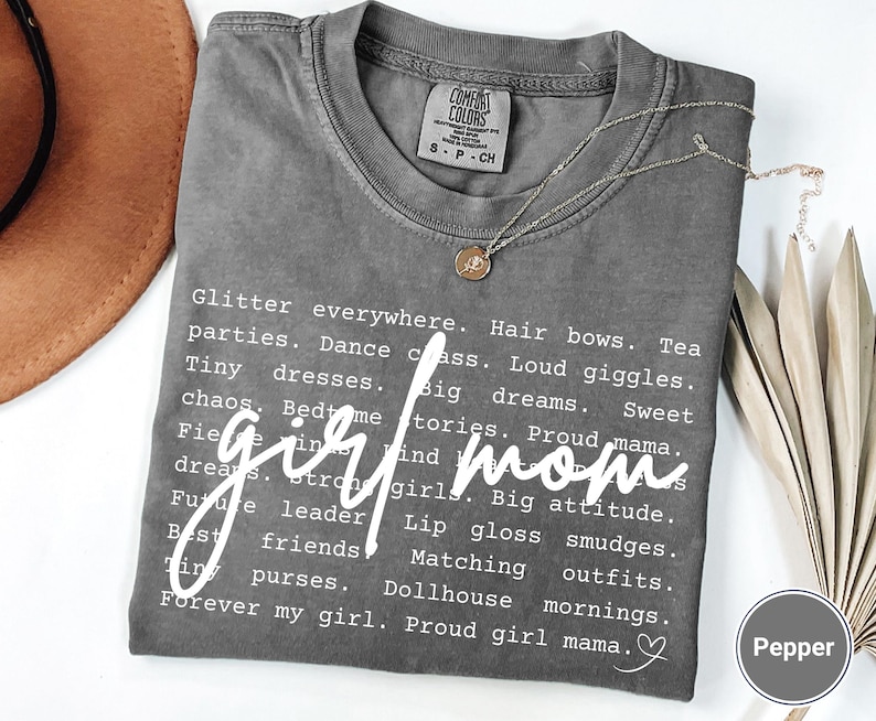 May include: A grey t-shirt with the words "girl mom" in a cursive font. The shirt features a list of related words in a white font. A gold necklace with a pendant is on the shirt.