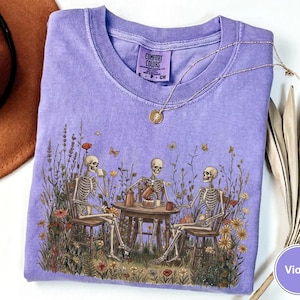 Może przedstawiać: Fioletowy t-shirt z kr&oacute;tkim rękawem z nadrukiem graficznym przedstawiającym trzy szkielety siedzące przy okrągłym stole na polu dzikich kwiat&oacute;w i pijące z kubk&oacute;w. Koszulka ma okrągły dekolt i metkę marki Comfort Colors.