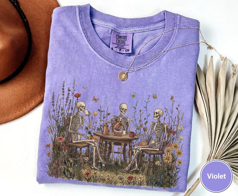 Może przedstawiać: Fioletowy t-shirt z kr&oacute;tkim rękawem z nadrukiem graficznym przedstawiającym trzy szkielety siedzące przy okrągłym stole na polu dzikich kwiat&oacute;w i pijące z kubk&oacute;w. Koszulka ma okrągły dekolt i metkę marki Comfort Colors.