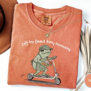 May include: A yam-colored t-shirt with the phrase "off to feed tiny humans" printed in white. The shirt features a cartoon frog riding a scooter and carrying a tray of food. A gold necklace is draped across the shirt.
