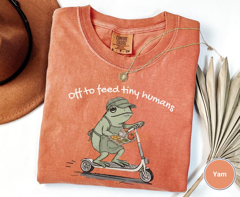 May include: A yam-colored t-shirt with the phrase "off to feed tiny humans" printed in white. The shirt features a cartoon frog riding a scooter and carrying a tray of food. A gold necklace is draped across the shirt.