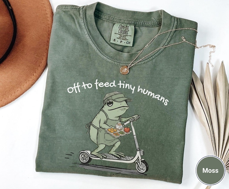May include: Moss green t-shirt with the phrase "off to feed tiny humans" and a cartoon frog riding a scooter, holding a tray of food. The shirt has a crew neck and short sleeves. A gold necklace is draped across the shirt.