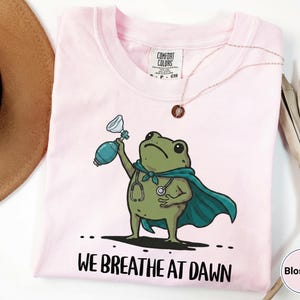 Pode incluir: Uma t-shirt rosa claro com um gr&aacute;fico de um sapo verde vestindo uma capa azul-petr&oacute;leo e um estetosc&oacute;pio, segurando um bal&atilde;o de reanima&ccedil;&atilde;o manual. Abaixo do sapo, o texto diz WE BREATHE AT DAWN. A t-shirt tem uma etiqueta Comfort Colors.