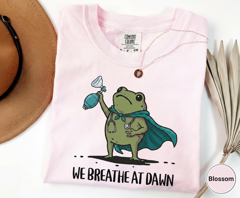 Pode incluir: Uma t-shirt rosa claro com um gr&aacute;fico de um sapo verde vestindo uma capa azul-petr&oacute;leo e um estetosc&oacute;pio, segurando um bal&atilde;o de reanima&ccedil;&atilde;o manual. Abaixo do sapo, o texto diz WE BREATHE AT DAWN. A t-shirt tem uma etiqueta Comfort Colors.