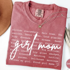 May include: Crimson t-shirt with the words "girl mom" in a cursive font. The shirt features a list of words associated with motherhood in a white font. A silver necklace with a pendant is draped across the shirt.