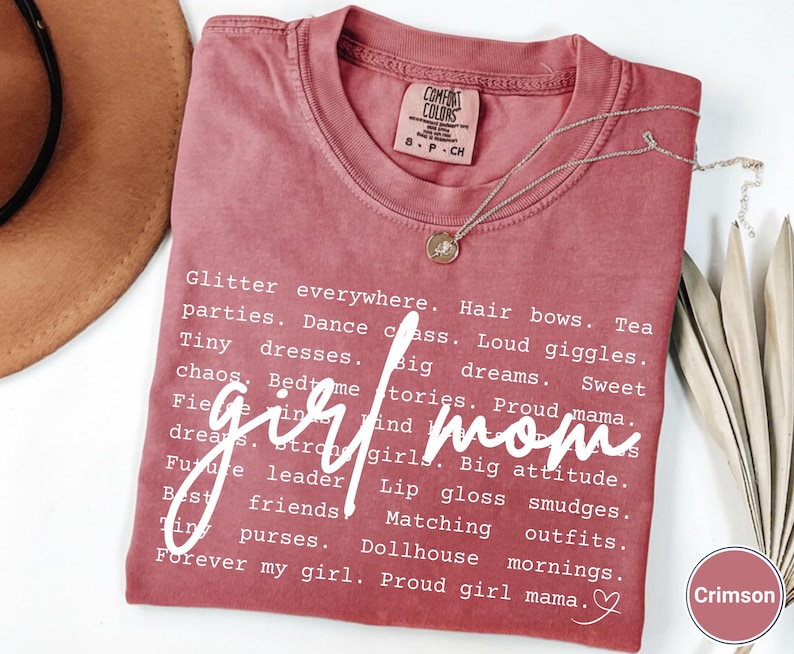 May include: Crimson t-shirt with the words "girl mom" in a cursive font. The shirt features a list of words associated with motherhood in a white font. A silver necklace with a pendant is draped across the shirt.