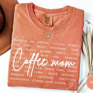 May include: A yam-colored Comfort Colors t-shirt with the words "Coffee mom" in a cursive font. The shirt has a list of coffee-related phrases in a white font. A gold necklace with a pendant is draped on the shirt.