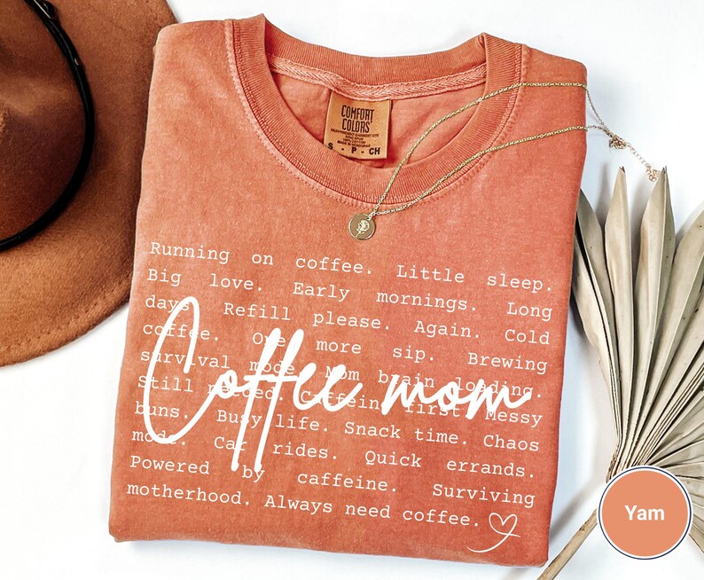 May include: A yam-colored Comfort Colors t-shirt with the words "Coffee mom" in a cursive font. The shirt has a list of coffee-related phrases in a white font. A gold necklace with a pendant is draped on the shirt.