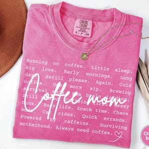 May include: A pink Comfort Colors t-shirt with the words "Coffee mom" in a cursive font. The shirt has a list of coffee-related phrases in white text. A gold necklace and a brown hat are also visible.