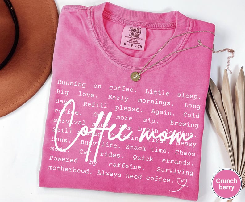 May include: A pink Comfort Colors t-shirt with the words "Coffee mom" in a cursive font. The shirt has a list of coffee-related phrases in white text. A gold necklace and a brown hat are also visible.