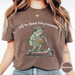 May include: Espresso-colored t-shirt featuring a cartoon frog on a scooter, holding a tray of food. The text "off to feed tiny humans" is arched above the frog. The frog wears a hat and apron. The shirt has a crew neck.