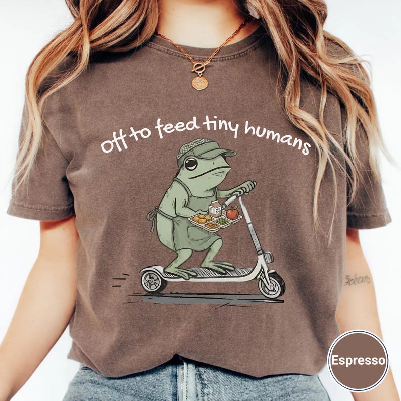 May include: Espresso-colored t-shirt featuring a cartoon frog on a scooter, holding a tray of food. The text "off to feed tiny humans" is arched above the frog. The frog wears a hat and apron. The shirt has a crew neck.