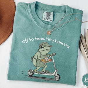 May include: Light green Comfort Colors t-shirt with the phrase "off to feed tiny humans" and a frog graphic riding a scooter. The frog is wearing a hat and apron and carrying a tray of food. A silver necklace is draped across the shirt.