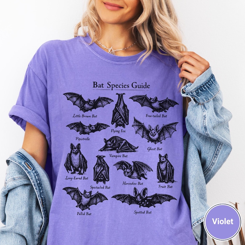 May include: A violet-colored t-shirt featuring a black graphic of various bat species with the text "Bat Species Guide." The shirt showcases illustrations of bats like the Little Brown Bat and the Flying Fox.