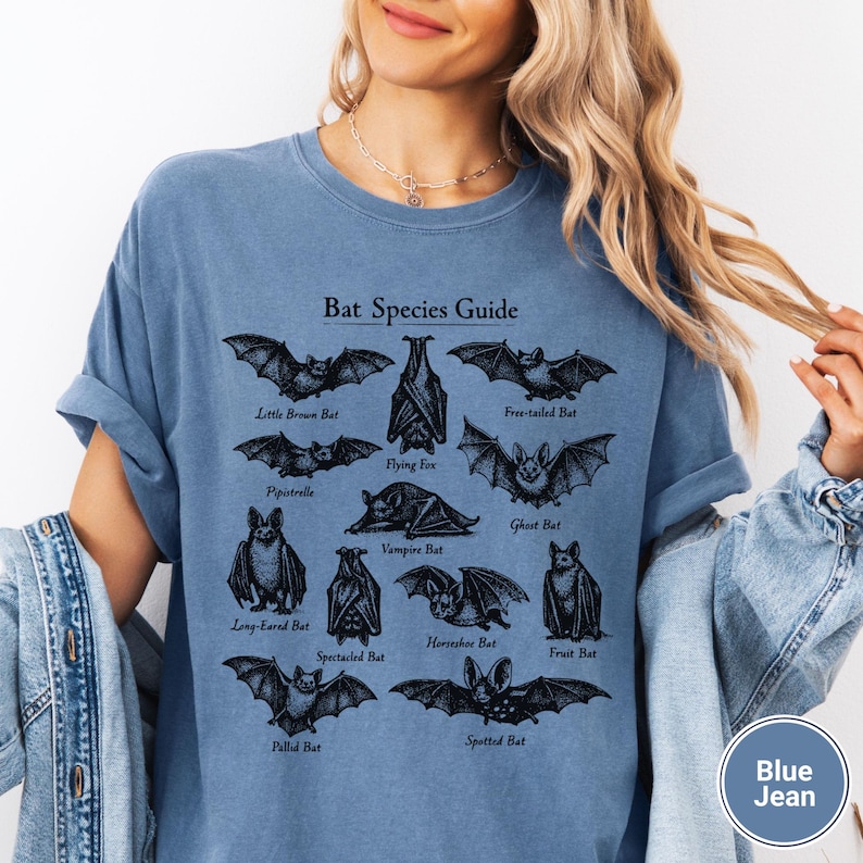 May include: Blue jean colored t-shirt featuring a black graphic of various bat species with the text "Bat Species Guide". The shirt has short sleeves and a relaxed fit, perfect for casual wear.