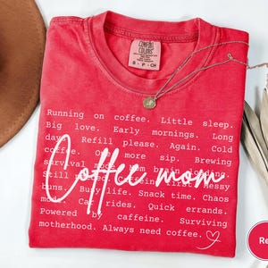 May include: Red Comfort Colors t-shirt with the words "Coffee Mom" in a cursive font. The shirt features a list of coffee-related phrases in white text. A gold necklace and a brown hat are also visible.