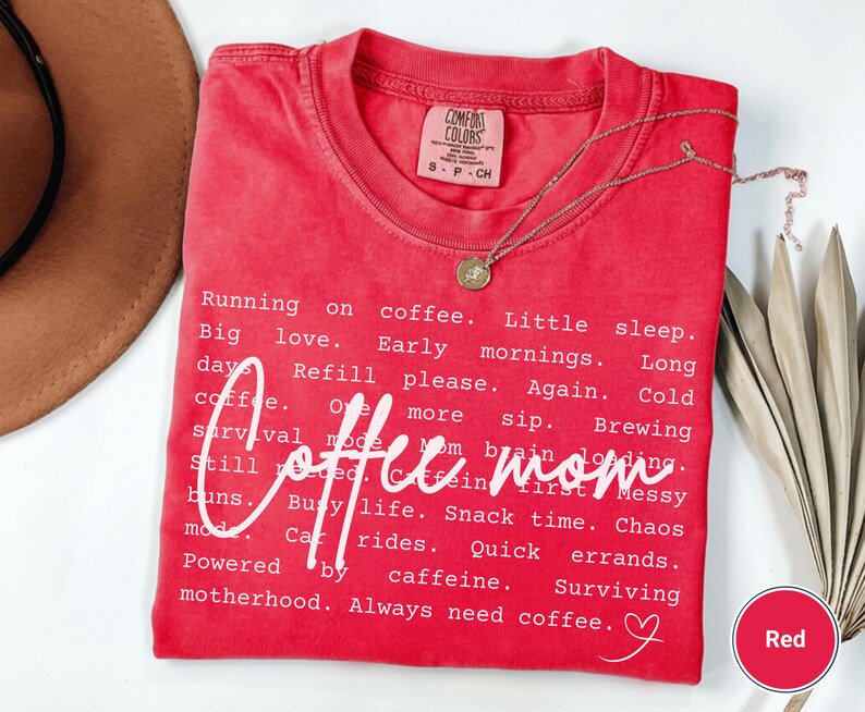 May include: Red Comfort Colors t-shirt with the words "Coffee Mom" in a cursive font. The shirt features a list of coffee-related phrases in white text. A gold necklace and a brown hat are also visible.