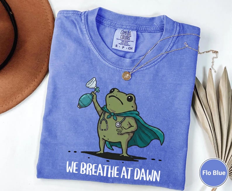 Pode incluir: Uma t-shirt azul pervinca de gola redonda com um gr&aacute;fico de um sapo verde usando uma capa verde-azulado e um estetosc&oacute;pio, segurando um ressuscitador manual. O texto "WE BREATHE AT DAWN" est&aacute; impresso em branco abaixo do sapo. Etiqueta Comfort Colors.