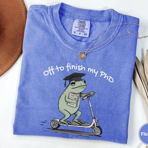 May include: Flo Blue t-shirt with the text "off to finish my PhD". The shirt features a cartoon frog wearing a graduation cap and riding a scooter. The frog is holding a book labeled "Draft Research".