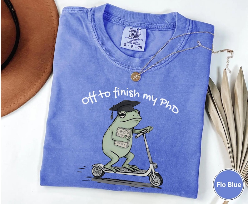 May include: Flo Blue t-shirt with the text "off to finish my PhD". The shirt features a cartoon frog wearing a graduation cap and riding a scooter. The frog is holding a book labeled "Draft Research".