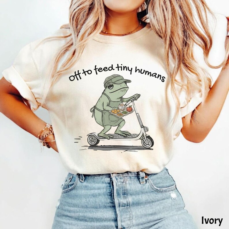 May include: Ivory-colored t-shirt with the text "off to feed tiny humans." The shirt features a cartoon frog riding a scooter and carrying a tray of food. The frog is wearing an apron and a cap. The shirt is made of a soft material.
