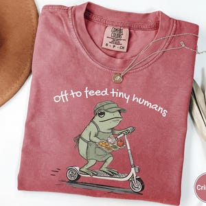 May include: Crimson-colored t-shirt with the text "off to feed tiny humans." The shirt features a cartoon frog riding a scooter and holding a tray of food. A silver necklace is draped across the shirt.