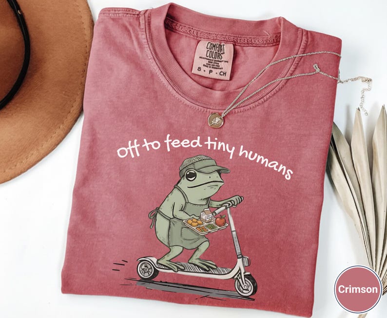 May include: Crimson-colored t-shirt with the text "off to feed tiny humans." The shirt features a cartoon frog riding a scooter and holding a tray of food. A silver necklace is draped across the shirt.