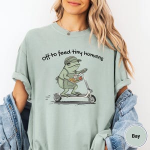 May include: A light green t-shirt with the phrase "off to feed tiny humans" printed in black. The shirt features a cartoon frog riding a scooter and carrying a tray of food. The frog is wearing a hat and apron.