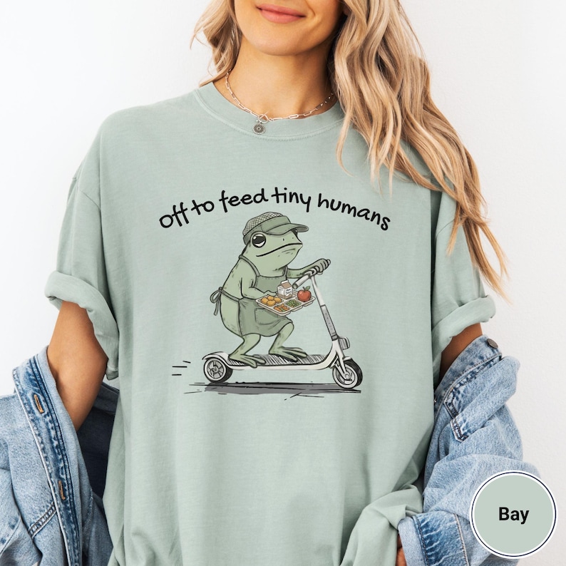 May include: A light green t-shirt with the phrase "off to feed tiny humans" printed in black. The shirt features a cartoon frog riding a scooter and carrying a tray of food. The frog is wearing a hat and apron.