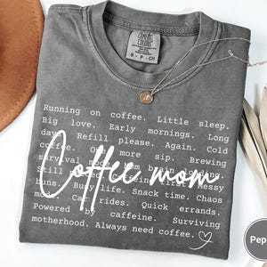 May include: A gray t-shirt with the words "Coffee mom" in a cursive font. The shirt has a list of coffee-related phrases. A gold necklace and a brown hat are also visible.