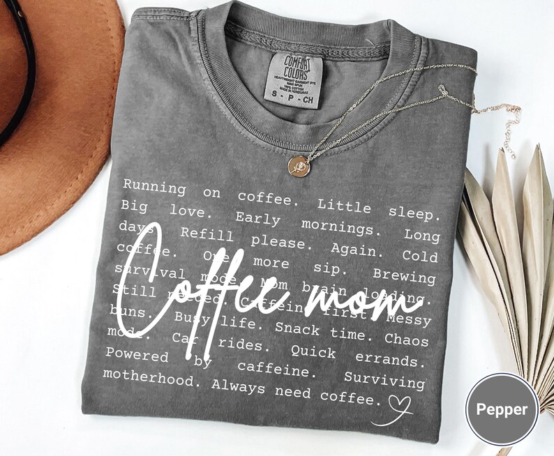 May include: A gray t-shirt with the words "Coffee mom" in a cursive font. The shirt has a list of coffee-related phrases. A gold necklace and a brown hat are also visible.