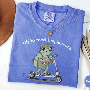 May include: Flo Blue t-shirt with the text "off to feed tiny humans." The graphic shows a frog wearing a hat and apron, riding a scooter while carrying a tray of food. The shirt has a crew neck and short sleeves.
