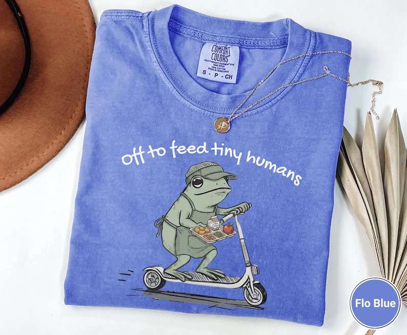 May include: Flo Blue t-shirt with the text "off to feed tiny humans." The graphic shows a frog wearing a hat and apron, riding a scooter while carrying a tray of food. The shirt has a crew neck and short sleeves.