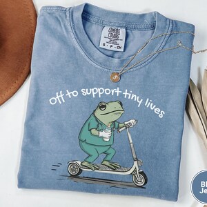 May include: Blue jean Comfort Colors t-shirt with the text "off to support tiny lives." The shirt features a cartoon frog wearing scrubs and riding a scooter. A gold necklace and a brown hat are also visible.