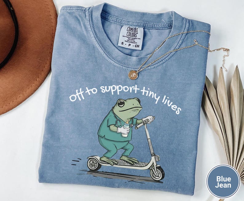 May include: Blue jean Comfort Colors t-shirt with the text "off to support tiny lives." The shirt features a cartoon frog wearing scrubs and riding a scooter. A gold necklace and a brown hat are also visible.
