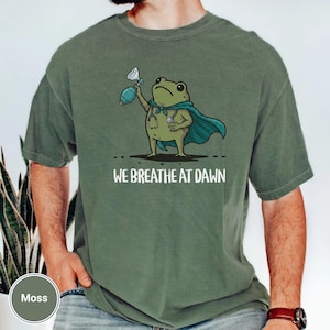Pode incluir: Uma t-shirt de manga curta verde musgo com o gr&aacute;fico de um sapo de desenho animado a usar uma capa azul-petr&oacute;leo e um estetosc&oacute;pio, segurando um ressuscitador manual. Abaixo, o texto WE BREATHE AT DAWN em letras brancas mai&uacute;sculas.