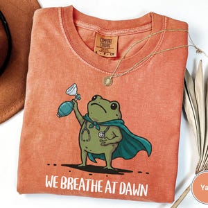 Pode incluir: Uma t-shirt cor de laranja queimado com o gr&aacute;fico de um sapo verde a usar uma capa azul-petr&oacute;leo e um estetosc&oacute;pio, segurando um bal&atilde;o de reanima&ccedil;&atilde;o. O texto "WE BREATHE AT DAWN" est&aacute; impresso por baixo. Etiqueta Comfort Colors vis&iacute;vel.