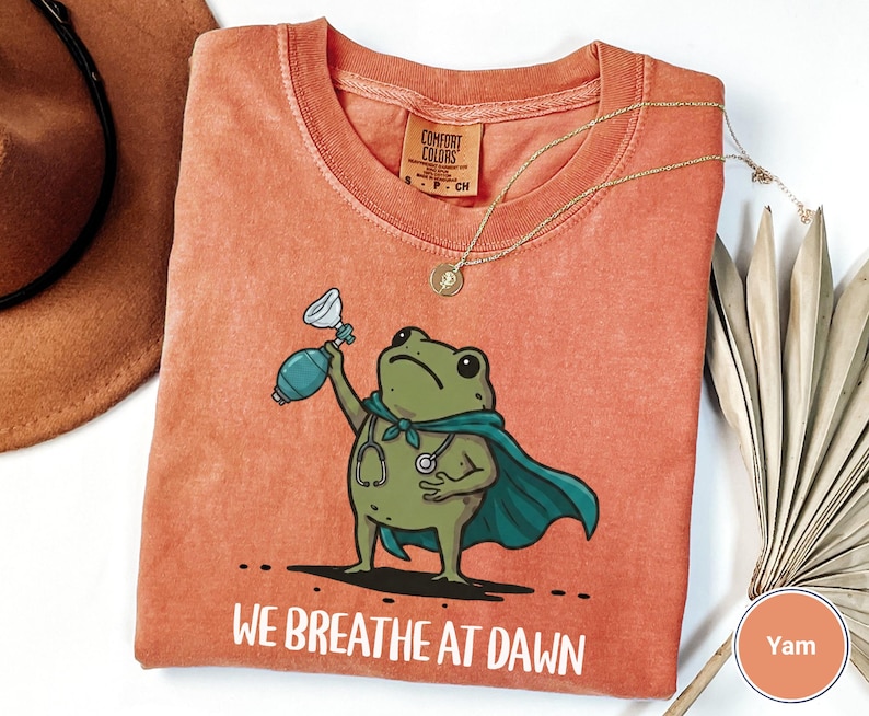 Pode incluir: Uma t-shirt cor de laranja queimado com o gr&aacute;fico de um sapo verde a usar uma capa azul-petr&oacute;leo e um estetosc&oacute;pio, segurando um bal&atilde;o de reanima&ccedil;&atilde;o. O texto "WE BREATHE AT DAWN" est&aacute; impresso por baixo. Etiqueta Comfort Colors vis&iacute;vel.