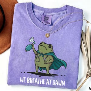 Pode incluir: Uma t-shirt violeta de manga curta com um gr&aacute;fico de um sapo verde a usar uma capa azul-petr&oacute;leo e um estetosc&oacute;pio, segurando um bal&atilde;o de reanima&ccedil;&atilde;o. O texto "WE BREATHE AT DAWN" est&aacute; impresso a branco por baixo do sapo.