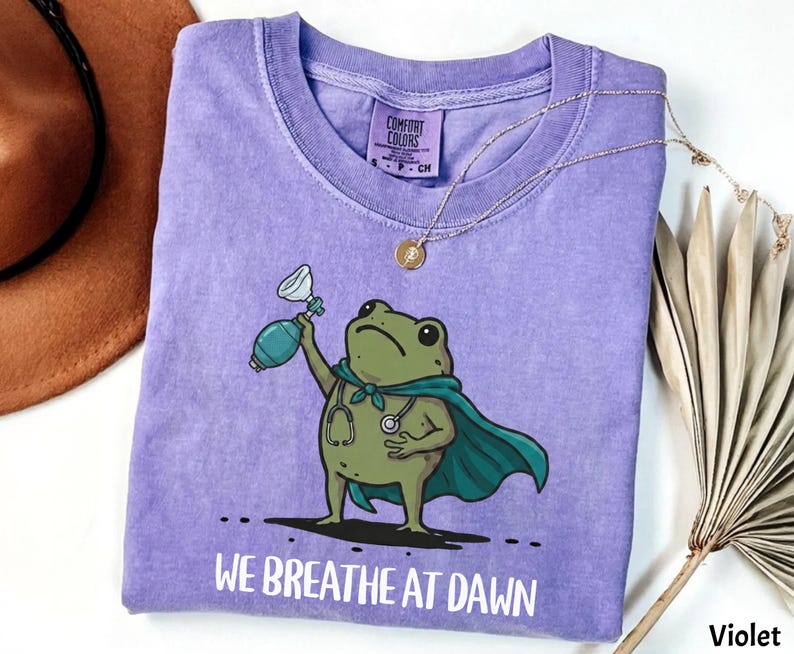 Pode incluir: Uma t-shirt violeta de manga curta com um gr&aacute;fico de um sapo verde a usar uma capa azul-petr&oacute;leo e um estetosc&oacute;pio, segurando um bal&atilde;o de reanima&ccedil;&atilde;o. O texto "WE BREATHE AT DAWN" est&aacute; impresso a branco por baixo do sapo.